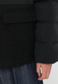 Melnā pūkera jaka ar tekstūru melnā auduma kabatu, piedāvā gludu ārpusi un nedaudz spīdīgu apdari.