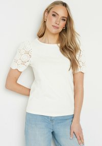 T-shirt en coton blanc avec des manches courtes en dentelle festonnée, texture lisse, encolure ras du cou, associé à un jean en denim bleu clair.