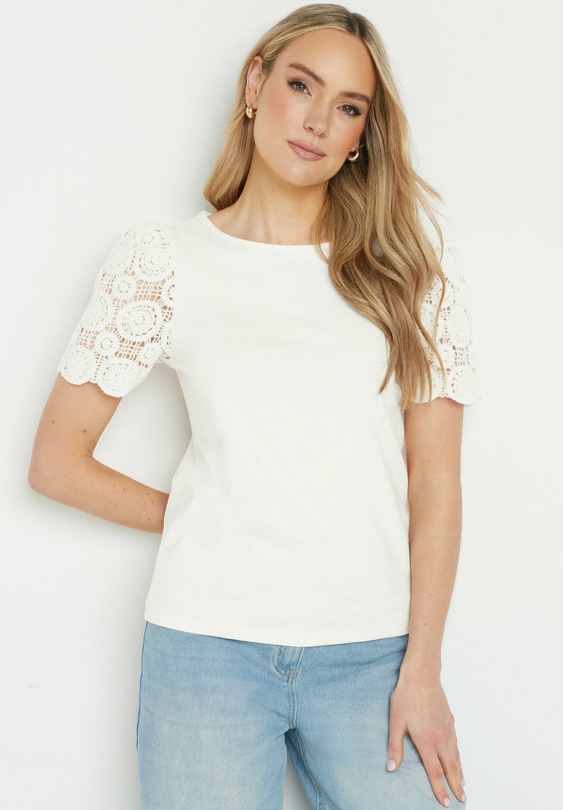 T-shirt en coton blanc avec des manches courtes en dentelle festonnée, texture lisse, encolure ras du cou, associé à un jean en denim bleu clair.