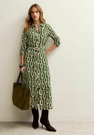 Groene jurk met een geometrisch patroon in het wit, knopen tot de borst, ceintuur om de taille en lange mouwen. Gecombineerd met een donkere suede tas en enkellaarsjes.
