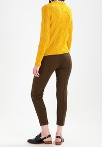 Gul stickad tröja med diamantstruktur, i kombination med bruna åtsittande byxor och svarta sandaler. Enkelt mönster, inga synliga mönster eller logotyper.