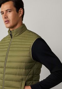 Olivgrüne, gesteppte Weste mit hohem Kragen, Reißverschluss vorne und ohne Ärmel, kombiniert mit einem schwarzen Langarmshirt darunter.