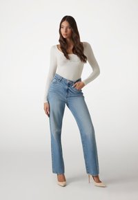 White long-sleeve bodysuit with a rounded neckline, paired with light blue high-waisted jeans and nude high heels. Smooth fabric and relaxed fit.