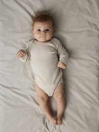 Šviesiai smėlio spalvos ribuotas kūdikio bodis ilgomis rankovėmis, su apvalia apykakle. Kūdikis guli ant šviesiai pilkos audinio paviršiaus, žiūrėdamas į viršų.