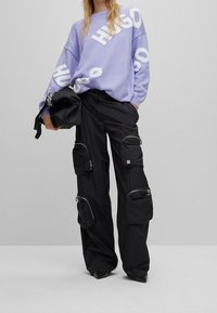 Lichtpaarse gebreide trui met witte "HUGO"-logo en zwarte cargobroek met meerdere ritssloten en een losse pasvorm. Zwarte handtas inbegrepen.