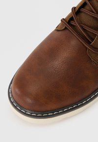 Zapato de cuero marrón con punta redondeada, parte superior texturizada, costuras blancas en contraste a lo largo de la suela y cordones marrones.