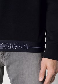 Svart långärmad skjorta i bomullsblandning, med ett kontrasterande marinblått band med vit broderad logotyp vid fållen. Grå denim är synlig.