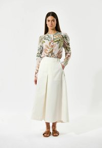 Blusa floreale a maniche lunghe con spalle a sbuffo in colori pastello, abbinata a una gonna bianca alla caviglia con pieghe frontali. Sandali marroni.