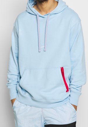 Homme portant un sweat à capuche bleu clair avec des cordons blancs et rouges et une poche avant avec une fermeture éclair rouge, mains dans les poches, vue recadrée.