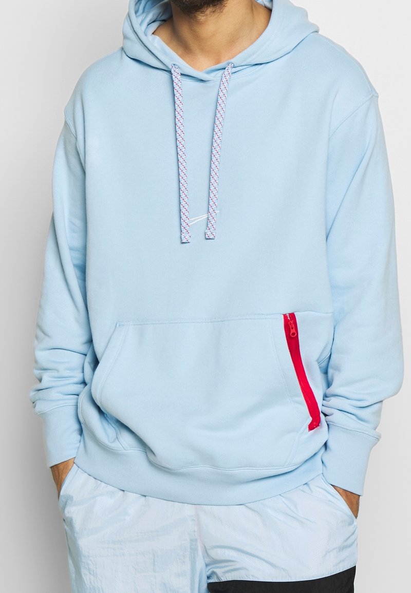Homme portant un sweat à capuche bleu clair avec des cordons blancs et rouges et une poche avant avec une fermeture éclair rouge, mains dans les poches, vue recadrée.