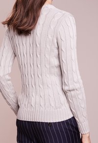 Pull en tricot gris clair avec un motif torsadé, ourlet côtelé et manches longues. Le fond est d'une couleur rose douce.