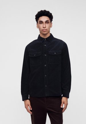 Jeune homme portant une chemise noire en velours côtelé boutonnée avec des poches poitrine, debout devant un fond gris clair uni.