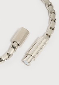 Ασημένιο βραχιόλι με μεταλλική αλυσίδα και διακοσμητικό κυλινδρικό κούμπωμα με χαραγμένα γεωμετρικά σχέδια και λείο, γυαλιστερό φινίρισμα.