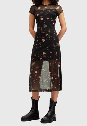 Femme portant une robe midi noire à imprimé floral avec des manches courtes transparentes et un jupon superposé, associée à des bottines noires épaisses.