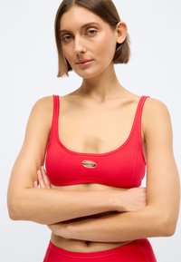 Γυναίκα με κοντά καστανά μαλλιά που φοράει κόκκινο αθλητικό σουτιέν με χρυσό λογότυπο, με σταυρωμένα τα χέρια, μπροστά σε απλό λευκό φόντο.