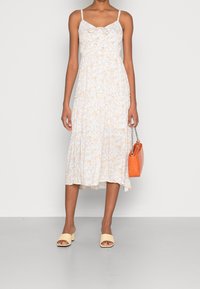 Weißes Blumen-Midikleid mit dünnen Trägern, Rüschenstufen und einer Schleifendetail am Dekolleté. Kombiniert mit beigen Blockabsatzsandalen und einer orangefarbenen Tasche.