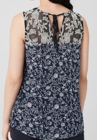 Granatowa bluzka w kwiaty z przezroczystym, haftowanym tyłem, z regulowanym zapięciem na wiązanie, miękkiego materiału i okrągłym dołem.