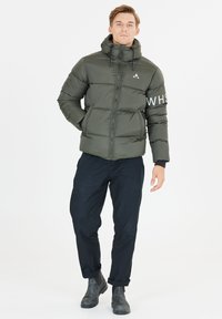 Olivgrön pufferjacka med huva, med horisontell sömnad, dragkedjestängning och vit text på ärmen. Matchas med svarta byxor och kängor.