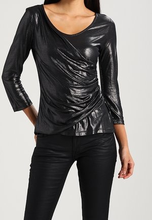 Femme portant un haut noir brillant à manches longues de style portefeuille avec des détails froncés, associé à un pantalon noir ajusté.