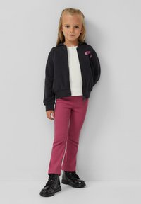 Zwarte zip-up hoodie met roze hart borduurwerk, witte lange mouwen top, roze ribbroek en glanzende zwarte enkellaarsjes.