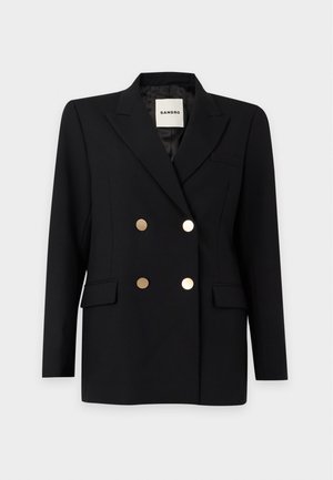 Kvinde står iført sort dobbeltradet blazer med guldknapper, sorte bukser med vide ben og en hvid blonder-top under.
