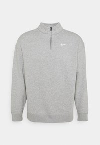Šedý fleecový pulovr s vysokým límcem, předním polozipskem a dlouhými rukávy. Na hrudi má malé bílé logo Nike. Jemná textura.