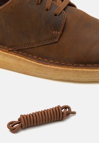 Brown leather shoe with a rounded toe, contrast stitching, and a light-colored sole. A brown shoelace is coiled next to it.