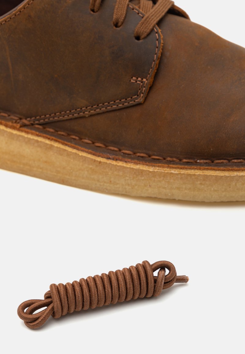 Zapato de cuero marrón con punta redondeada, costuras en contraste y suela de color claro. Un cordón marrón está enrollado al lado.