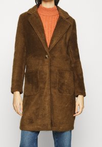 Hnědý kožešinový kabát s klopeným límcem, jednobotoním zapínáním a dvěma předními kapsami. Nosený přes oranžový svetr, v kombinaci s modrými džíny.