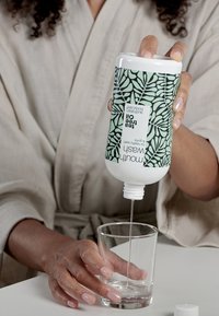 Weiße Kunststoffflasche mit grünem Blättermuster, die einen klaren Flüssigkeit in ein Glas gießt. Der Flaschenverschluss ist abgenommen, was einen Ausgießer zeigt.