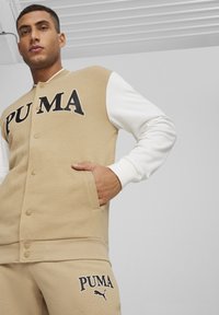 Felpa beige con maniche bianche, caratterizzata da un grande logo "PUMA" e bottoni a pressione. Il materiale sembra morbido con una texture opaca.