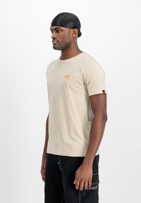 Beige T-Shirt mit kurzen Ärmeln und einem orangefarbenen Logo auf der Brust. Das Material wirkt weich und leicht, ideal für die Freizeit.