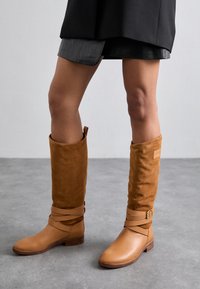 Bottes hautes jusqu'aux genoux en cuir et daim couleur tan, avec un bout arrondi, un talon plat et des sangles croisées ornées d'une boucle. Texture lisse, design minimaliste.