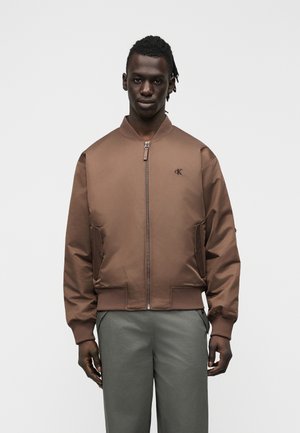 Mann trägt eine braune Bomberjacke mit Reißverschluss und "ck"-Logo auf der Brust, kombiniert mit grauer Hose, stehend vor einfachem weißem Hintergrund.