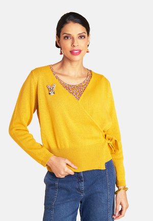 Frau trägt einen gelben Wickelpullover mit einer kleinen Tierbrosche, ein bunt gemustertes Oberteil, blaue Jeans und goldene Armbänder vor weißem Hintergrund.