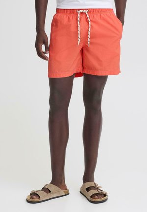 Mężczyzna w koralowych kąpielówkach z białym troczkiem, beżowych sandałach z klamrą, stojący na jednolitym jasnym tle.