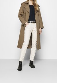 Beige trenchcoat med dobbeltknap-design, parret med hvide lige ben jeans og sorte ankelstøvler. Sort bælte med guldbuckle-detalje.