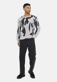 Un pull gris clair duveteux avec imprimé vache noir, un col rond et des manches longues, associé à un pantalon en denim noir et des chaussures noires.