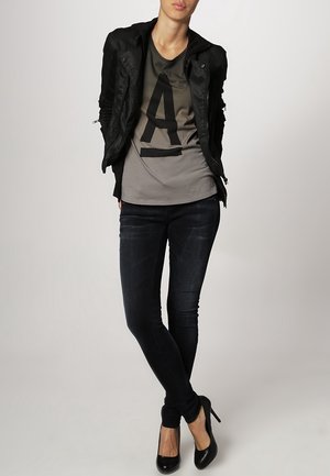 Zwarte korte jas, grijs T-shirt met een grote zwarte 'A', gecombineerd met donkere skinny jeans en zwarte hoge hakken. Eenvoudig en modern ontwerp.