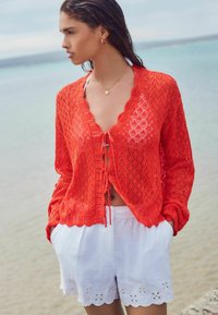 Cardigan en dentelle rouge tricoté avec des bords festonnés, noué à la taille ; associé à des shorts blancs avec des détails en œillet. Décor de plage.