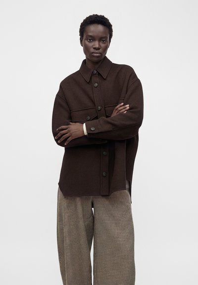 Personne aux cheveux courts et bouclés portant une veste marron foncé boutonnée et un pantalon ample beige à carreaux, debout les bras croisés.