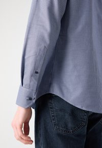Camicia a quadri blu e bianca con polsini abbottonati, caratterizzata da una texture morbida e orlo curvo, abbinata a jeans scuri.