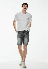 Bílé tričko s potiskem "Club Marina" na hrudi, spárované se šedými džínsovými šortkami a bílými sportovními teniskami s ohrnutými lemy.