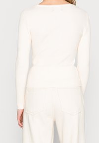 Ljusbeige ribbad långärmad topp med rundad halsringning, figurnära silhuett och subtila sömnadsdetaljer på baksidan.