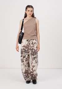 Asymmetrisk brun tank top parret med beigefargede og brune mønstrede vide bukser med snor i midjen. Modellen holder en svart veske.