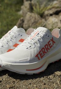Białe buty sportowe z pomarańczowym logo TERREX, siateczkową cholewką i amortyzowaną podeszwą, umieszczone na skalistym, zewnętrznym podłożu.