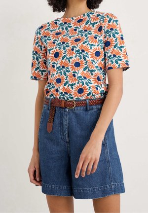 Kvinde iført orange og blå blomstret kortærmet skjorte med blå denimshorts og et brunt fletbælte.