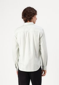 Chemise à manches longues vert clair avec des rayures blanches verticales, patte de boutonnage et ourlet arrondi. Le dos présente un pli pour plus de liberté de mouvement.