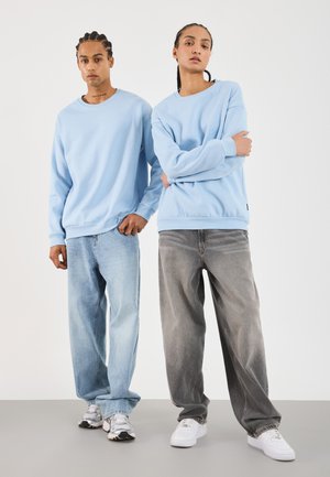 Twee mensen die naast elkaar staan, gekleed in lichtblauwe sweats, ontspannen jeans en sneakers tegen een effen witte achtergrond.