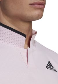 Polo rose clair avec un petit col noir, fabriqué dans un tissu texturé et arborant un logo Adidas sur le côté gauche.
